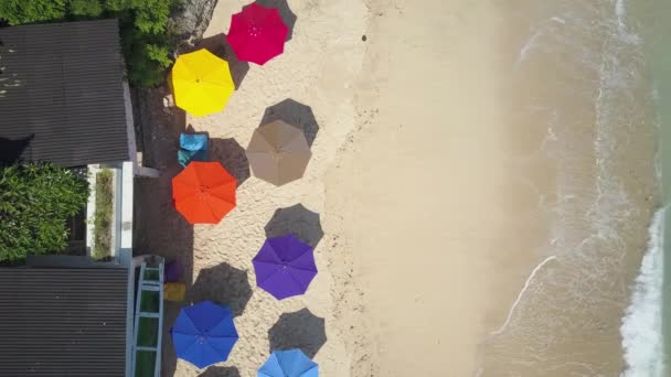 空中顶部 在美丽的空白色沙滩上 使用红色 黄色和蓝色的遮阳伞 海滨度假屋在阳光明媚的巴厘岛 海浪在海岸上沐浴着五颜六色的遮阳伞 — 图库视频影像