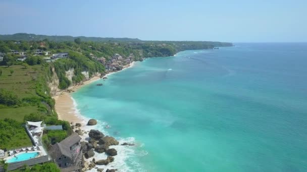 空中飞行 飞越郁郁葱葱的绿色岩壁海岸线 在令人惊叹的巴厘岛 印度尼西亚的深蓝色海洋之上升起 海边豪华旅游胜地的平房和阁楼与无限水池 — 图库视频影像