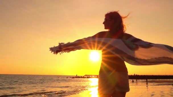 慢动作 镜头耀斑 迷人的年轻女子与太阳镜旋转在海滩上挥舞着挥舞着围巾在多风的夏夜在梦幻般的巴厘岛岛华丽的金色日落 — 图库视频影像
