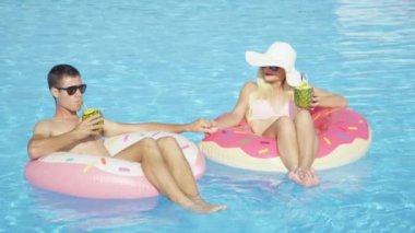 Ağır çekim: ananaslı kokteyl içme ve eğlence pembe halka floaties havuzunda rahatlatıcı genç çift. Genç adam ve kadın aşık şişme halka yüzen güneşli yaz tatil üzerinde rahatlatıcı