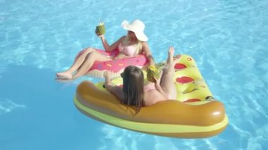 Ağır çekim yakın çekim: eğlenceye döşeme iki gülümseyen kız arkadaşım renkli floaties, yaz tatili alkollü içecekler içme. Kokteyl yudumlarken, şişme pizza ve pembe çörek üzerinde rahatlatıcı genç kadınlar