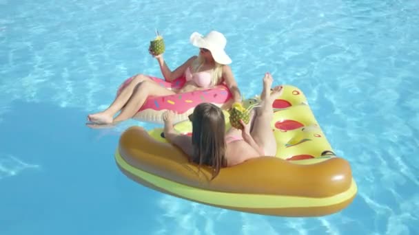 慢动作特写 两个面带微笑的女朋友躺在有趣的五颜六色的浮雕上 在暑假喝酒精饮料 年轻女性喝鸡尾酒 放松在充气比萨饼和粉红色甜甜圈 — 图库视频影像