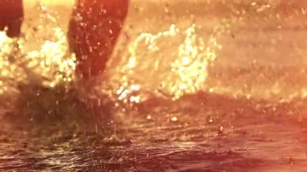 慢动作特写镜头耀斑 活跃的年轻人在金色日落时在沙滩上泼水 在美丽的黄色早晨日出时 无法辨认的男跑步者在浅海水域慢跑 — 图库视频影像