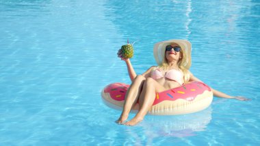 Yaz tatilinde, havuzda şişme donut şamandırasına uzanmış ananas alkollü içecek içen mutlu kadını yakından çek. Gülümseyen kız şişme yastıkta yüzüyor, kokteyl yudumluyor