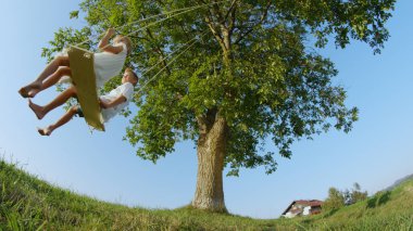 Düşük açı gülümseyen erkek ve kız eğleniyor büyük bir yeşil ağaç altında ahşap bir salıncak üzerinde sallanan. Mutlu iki genç çocuk bir ahşap sallanan set salıncak. Yaz çayır bir salıncak üzerinde zevk kardeşler