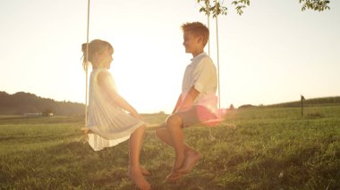 Lens Flare gülümseyen ve altın günbatımında eğleniyor ahşap salıncak üzerinde oturan kız. İki oyuncu çocuk her salıncakta konuşmaya karşı karşıya. Neşeli erkek ve kız yaz akşam zevk.