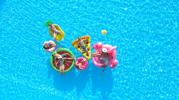 空中コピースペース 遊び心のある女の子と男がプールでカラフルな浮遊物に横たわっています 夏休みを楽しむ幸せな笑顔の友人インフレータブルパイナップル フラミンゴ スイカ ドーナツフロート — ストック写真