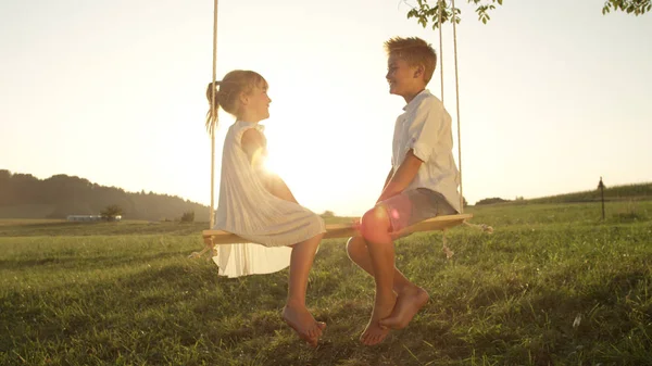 Lense 耀斑微笑的兄弟和姐妹坐在木秋千上 在金色的日落中享受乐趣 一对顽皮的孩子面对面地在秋千上说话 愉快的男孩和女孩享受夏天晚上 — 图库照片