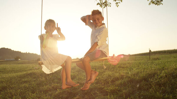 CLOSE UP: Smiling brother and sister playfully posing for the camera. Young siblings having fun and enjoying a warm summer evening. Youthful children sitting and playing on a swing at golden sunset