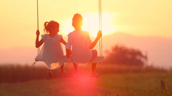 LENSE FLARE Adorable siblings hold hands swaying on late summer evening. Little brother and sister swinging on wood swing in meadow gazing at warm golden sunset. Young love blooming at gold sundown