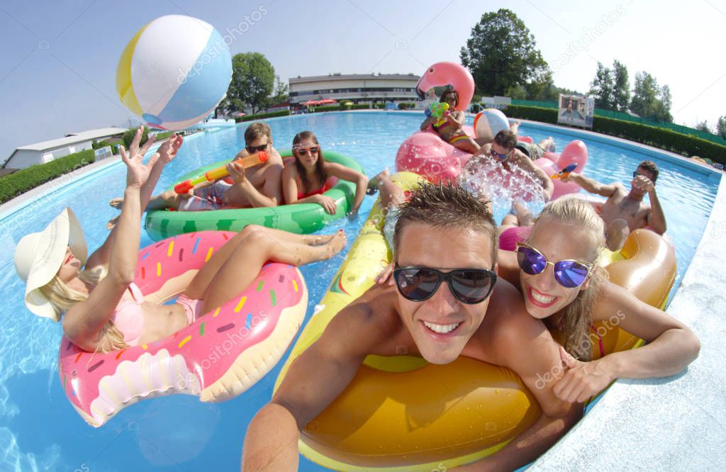SELFIE: Smiling young people enjoying on fun colorful floaties, playing volleyball at pool party. Happy teenagers on inflatable pizza, flamingo, watermelon and doughnut floats having watergun fight