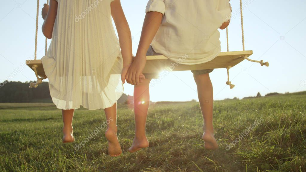 CLOSEUP LENSE FLARE Unrecognizable kids sitting on big wooden swing in sunny evening. Barefoot little brother and sister swinging in golden summer . Young love between boy and girl on romantic evening