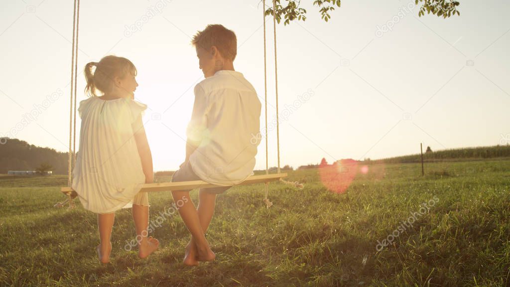 COPY SPACE LENSE FLARE Couple young kids cheerfully chatting and sitting on wood swing at gold sunset. Happy little girl and boy having fun on summer evening. Playful brother and sister talking.