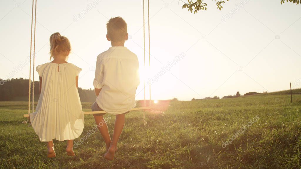 COPY SPACE LENSE FLARE: Two young children sitting on wooden swing gazing at the sunset. Little siblings enjoying a golden summer evening. Blooming love between boy and girl swinging on a swing set.