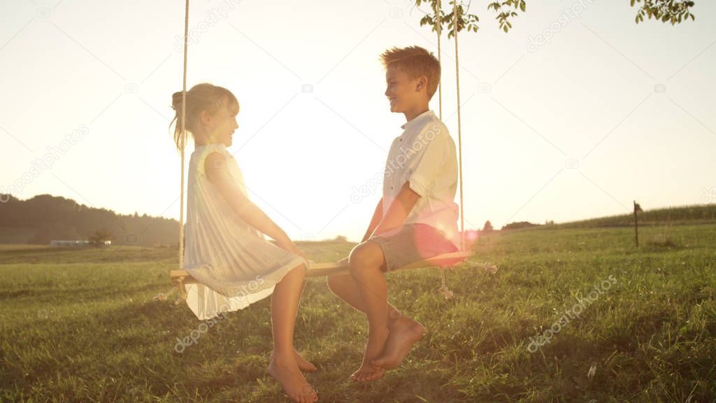 LENSE FLARE Smiling brother and sister sitting on wooden swing having fun at golden sunset. Couple of playful kids facing each other talking on swing. Cheerful boy and girl enjoying summer evening.