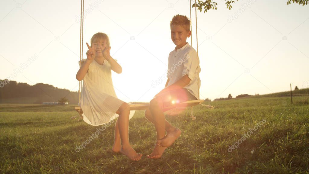 LENSE FLARE: Smiling brother and sister playfully posing for the camera. Young siblings having fun and enjoying a warm summer evening. Youthful children sitting and playing on a swing at golden sunset