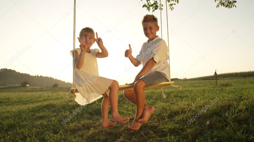 CLOSE UP: Smiling brother and sister playfully posing for the camera. Young siblings having fun and enjoying a warm summer evening. Youthful children sitting and playing on a swing at golden sunset
