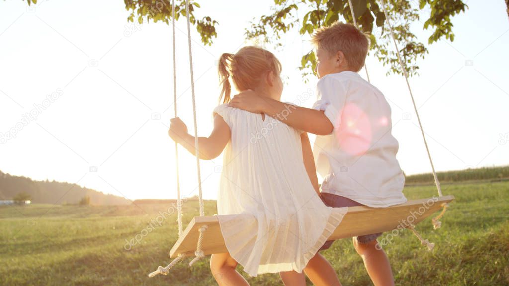 CLOSE UP LENS FLARE Couple children embracing on wooden swing at gold sunset. Young love blooming between boy and girl swinging on swing set. Brother hugging his little sister enjoying a warm evening