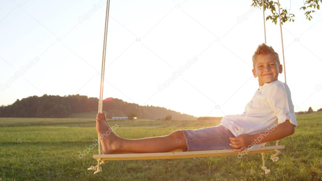 CLOSE UP: Smiling young boy relaxing on wooden swing posing for the camera. Laidback barefoot kid enjoying a golden sunset. Youngster unwinding on a warm summer evening sitting on swing under a tree