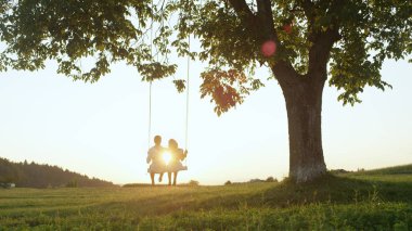 LENS FLARE SILHOUETTE: Young couple in love swaying on tree swing at golden summer sunset. Happy man and woman swinging under tree in sunny evening. Boyfriend and girlfriend hugging on romantic date. clipart