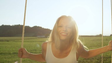 Mercek Parlaması, portre: doğada yaz güneş ışınları içinde sallanır gibi parlayan altın rengi ile mutlu genç kadın. Şakacı sıcak bahar kırsal kesimde sallanan beyaz sundress Bayan Kırmızı dudaklar ile gülüyor.