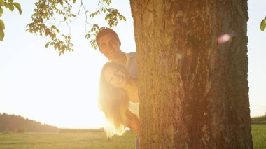 Yakın Lens Flare Up: bir yaz akşamı bir ağacın arkasından bakmak çift gülümsüyor. Genç erkek ve kadın bir ağaç gövdesi saklanıyor geçmiş parlayan yapraklara. Oğlan ve kız altın gün batımında romantik tarihte