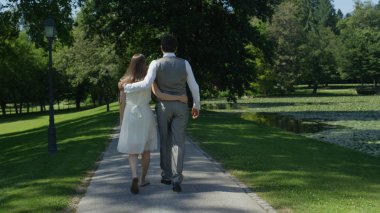 Back View: Sevgi dolu kocası yeşil parkta gezinen güzel hamile eşi kucaklayan. Genç mutlu çift bahçede rahatlatıcı yürüyüş sarılma. Aşık ebeveynlerin birbirini tutmalarını bekliyoruz.