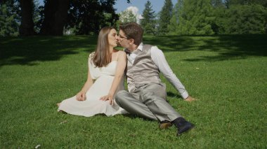 Güzel genç çift eğlenceli öpüşme güzel bir güneşli parkta çim üzerinde oturup. Anne-babalar sıcak güneşli bir günde bazı hassasiyet paylaşmak bekliyoruz. Yeni evliler yaz aylarında açık havada harika bir zaman yaşıyorsanız.