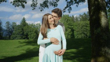 Kucaklanan hamile çift güneşli bir günde bir ağacın altında durarak samimi bir an paylaşıyor. Heyecanlı genç anne-babalar hamilelik harikaları zevk olmak. Karı koca bebeğin doğmasını bekleyemez.