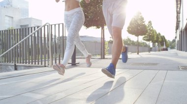 LOW ANGLE, LENS FLARE: Fit young couple jogging outdoors down asphalt walkway. Unrecognizable sporty couple jogging on a warm sunny morning. Fit Caucasian male and female training for marathon race.