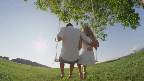 在阳光明媚的夏日里 无忧无虑的年轻夫妇在木绳秋千上摇曳 无法辨认的男友拥抱女朋友 而在金色的阳光下摇摆在一棵树在农村的环境 — 图库照片
