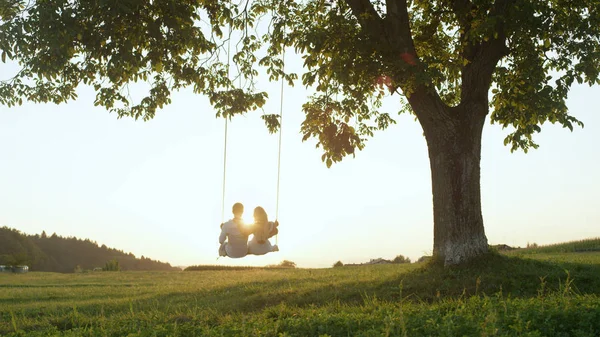 镜头闪烁剪影 设置阳光照耀在年轻夫妇的肩膀上的绳索秋千 在浪漫的夏季约会中 年轻的成年人靠在树下的秋千上 看着金色的日落 — 图库照片