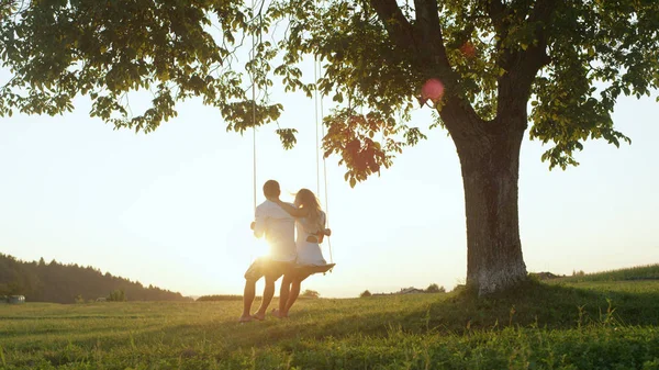 镜头闪烁剪影 可爱的年轻夫妇坐在金色的暮色下 在草甸的大树下的绳索秋千 在夏日的夜晚 男朋友和女朋友享受着约会 — 图库照片