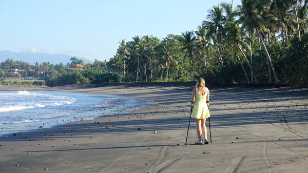 Unrecognizable young woman supporting herself on crutches after injuring her ankle by stepping on a pebble in flip flops Young lady looking down the lovely sandy shore in remorse after hurting herself