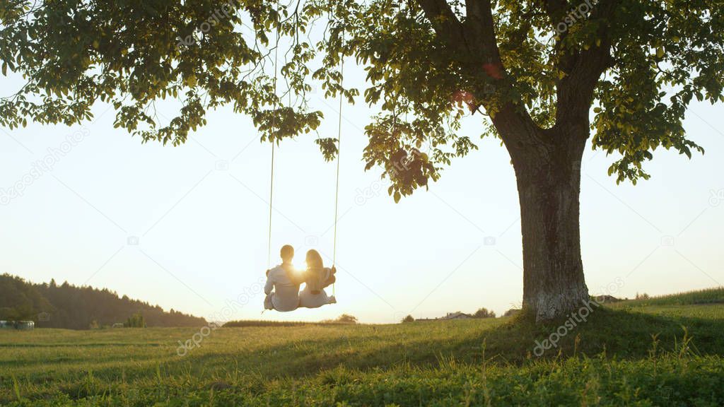 LENS FLARE SILHOUETTE: Setting sun shining over young couple's shoulders on rope swing. Young adults on romantic summer date, leaning back embraced on swing under tree and watching the golden sunset