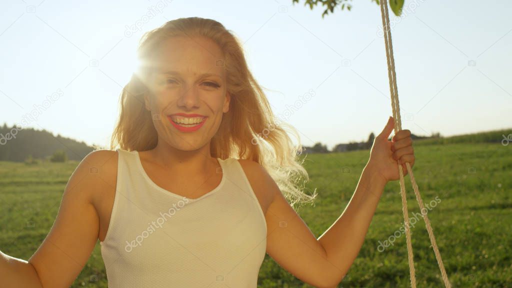 LENS FLARE PORTRAIT: Happy young woman in white sundress sways in sunny nature. Wavy golden hair flying in warm summer air. Joyful young female with blonde hair enjoying warm springtime on rope swing.