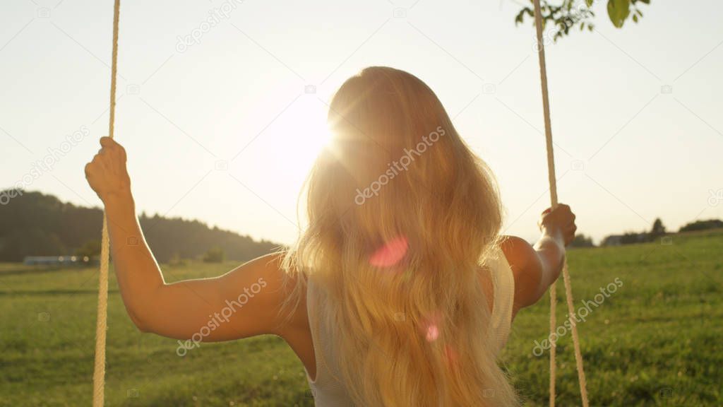 LENS FLARE, CLOSE UP: Unrecognizable young woman with long blonde hair looking into beautiful spring sunset while swinging in meadow. Thoughtful lonely girl swaying in nature, glowing in spring sun.