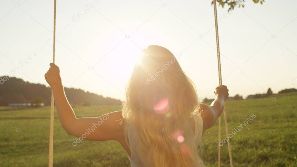 LENS FLARE, CLOSE UP: Unrecognizable woman in white gazing into distance at sunset. Young unknown female wrapped in thought as she sways on rope swing. Twilight sunbeams shining on young lady on swing