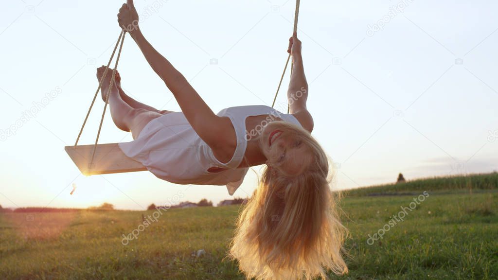 LENS FLARE CLOSE UP: Happy young blonde woman in white dress looking back and smiling into the camera while swinging in golden summer sunset in country meadow. Cheerful lady leaning back in her swing