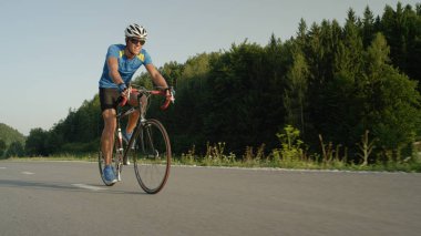 Düşük açılı: Genç erkek motorcu güneşli yaz öğleden sonra yarış sırasında boş asfalt yolda pedal. Uzun mesafe Tur ile yeşil kırsal manzara yolculuk zevk eğlence bisikletçi.