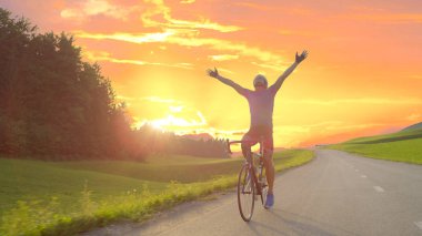 LENS FLARE: Thrilled bicycle rider celebrates win in bike race across countryside by outstretching arms and looking into the golden evening sky. Pro road cyclist rejoicing after completing workout. clipart