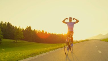 Mercek Parlaması, kopya alanı: Muzaffer yol motorcu altın yaktı doğada zorlu yarışı bitirmek mutlu. Motosiklet gidonun gidelim ve onun kazanmak yol bisiklet yarışı içinde kutlamak için kollarını outstretches.