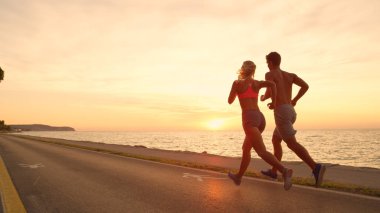 Kopya alanı: Tanınmayan genç çift günbatımında çarpıcı okyanus yolda jogs. Güzel akşam altın ışığı aşağı atletik erkek ve kadın üzerinde etkin tarih Okyanusu yakınlarındaki parlar. Muhteşem sahil gündoğumu