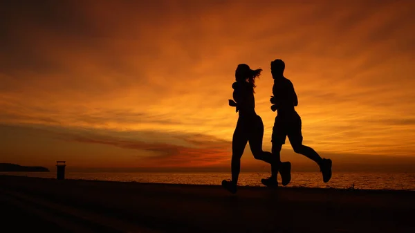 复制空间 剪影年轻的男人和女人做他们的晚上锻炼在宁静的海水前在华丽的日落 在橙色日落时沿着海洋慢跑的无法辨认的情侣的完美镜头 — 图库照片