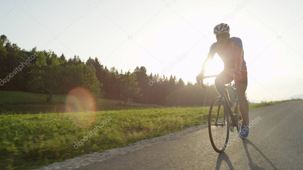 COPY SPACE, LENS FLARE: Professional male road biker enjoying a sunny cruise on his bicycle. Active man with sunglasses racing in a professional road bicycle race through sunny spring countryside.