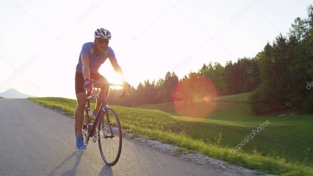 LENS FLARE: Sporty cyclist on road bike training on a sunny day in pretty green nature. Athletically built Caucasian male with sunglasses rides his road bicycle through beautiful spring landscape.