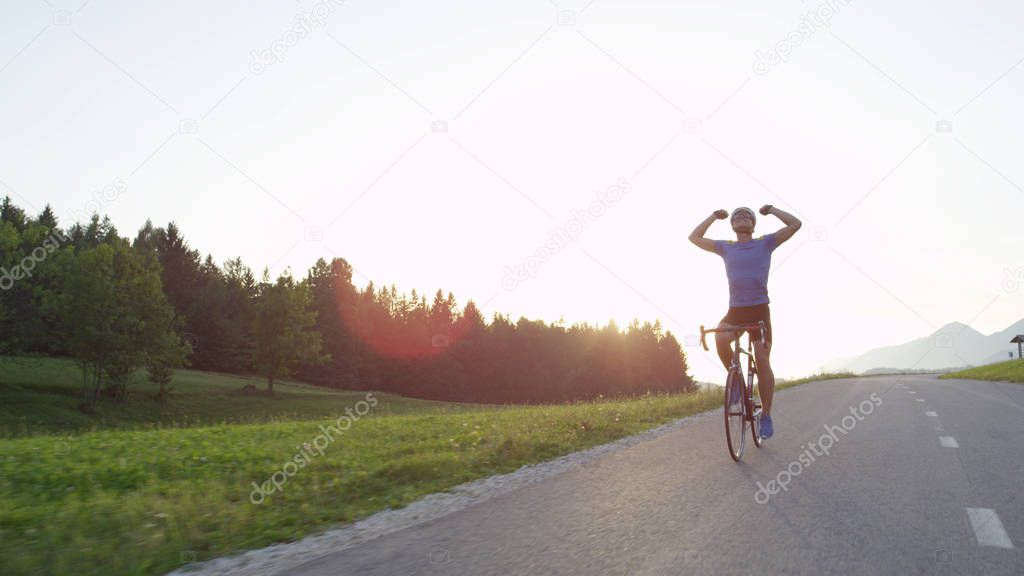 SUN FLARE: COPY SPACE: Thrilled road biker relieved to have finished his intense workout in the golden lit countryside. Happy young athlete pumping his fists in the air and cruising on his bicycle.