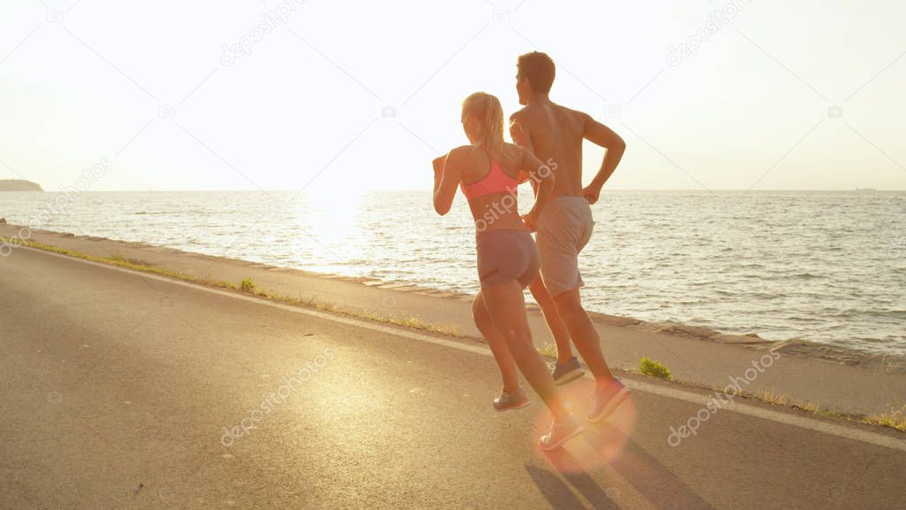 SUN FLARE: Lovely young Caucasian couple jogs down asphalt ocean road on perfect summer afternoon. Unknown active female and male exercising in the summer sun rays near peaceful beach during holiday.