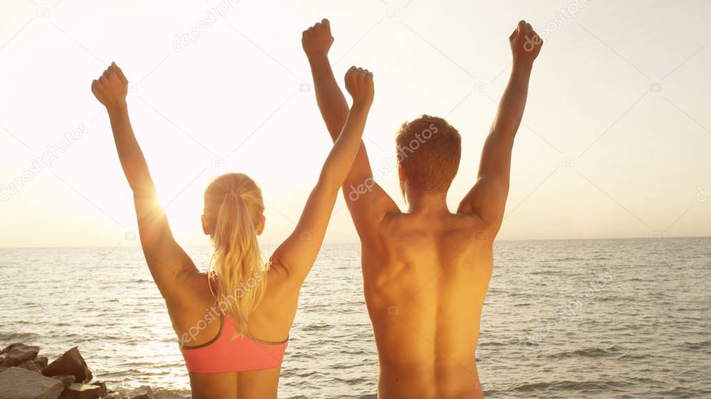 CLOSE UP, SUN FLARE: Joyous young couple outstretch their arms after fun run at the seaside. Unrecognizable fit man and woman lift their arms overhead in celebration while looking at the sunny ocean.