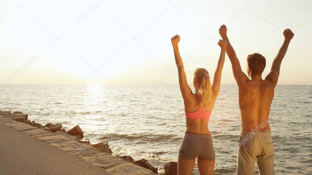 CLOSE UP, COPY SPACE: Young fit couple finishes scenic run at the seaside and raises their arms. Athletic boy and girl celebrate successful run near rocky beach on perfect day in summer.